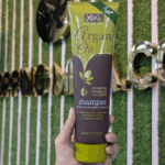 Hand holding a tube of XHC Argan Oil Shampoo in front of a green textured wall. The packaging features green and brown colors and highlights its hydrating, nourishing, and cleansing formula with Moroccan argan oil extracts.