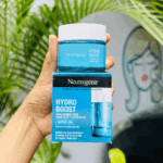 A hand holding Neutrogena Hydro Boost Water Gel 50ml above its retail box, surrounded by green leaves and a soft wall mural in the background. The packaging highlights Hyaluronic Acid, Amino Acids, and Electrolytes for deep hydration and smooth, dewy skin.