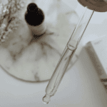 Close-up of The Ordinary Multi-Peptide Serum dropper releasing a clear liquid, showing the serum’s lightweight consistency. The blurred background includes the product bottle and box on a marble surface.