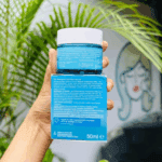 The back view of Neutrogena Hydro Boost Aqua Gel 50ml jar and box held in hand, showing product details and ingredients list. The background features lush green plants and a wall mural, emphasizing a clean, hydrating, and refreshing skincare aesthetic.