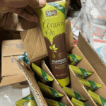 A hand holding a tube of XHC Argan Oil Shampoo from a cardboard box filled with multiple units. The green and brown packaging highlights its vegan, hydrating, nourishing, and cleansing formula with Moroccan argan oil extracts.