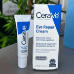 CeraVe Eye Repair Cream tube and box placed on a table with a leafy background, showing the product’s blue-and-white packaging that highlights its benefits for dark circles and puffiness.