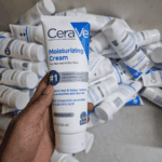 Hand holding CeraVe Moisturizing Cream tube in front of bulk stock showing normal to dry skin moisturizer packaging