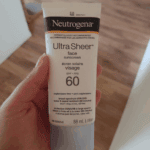 Close-up of a hand holding Neutrogena Ultra Sheer face sunscreen SPF 60 (88ml) tube indoors with wooden floor background.