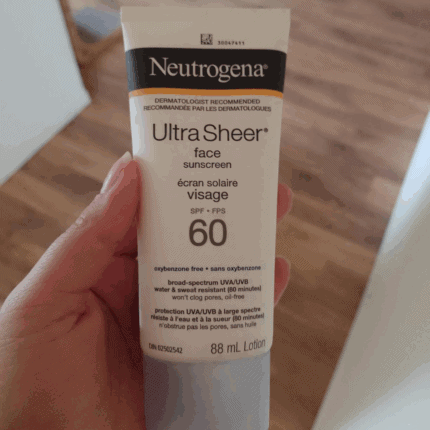 Close-up of a hand holding Neutrogena Ultra Sheer face sunscreen SPF 60 (88ml) tube indoors with wooden floor background.