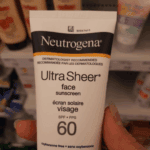 Hand holding Neutrogena Ultra Sheer face sunscreen SPF 60 (88ml) tube in a Skinify store aisle with skincare products on shelves in the background.