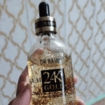 Hand holding a Dr Rashel 24K Gold Radiance and Anti-Aging Primer Serum bottle with dropper, showing suspended gold flakes inside the clear serum, photographed indoors against a patterned wall background.