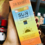 Close-up of a hand holding the Dr. Rashel Sun Cream Whitening SPF 75 box with gradient orange, yellow, and blue packaging in a store setting.