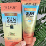 Hand holding Dr. Rashel Sun Cream Whitening SPF 75 tube alongside its box with gradient orange, yellow, and blue packaging against green leaves background.