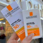 Hand holding the sunscreen bottle and box with white and orange packaging in a blurred store background.