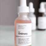 The Ordinary Lactic Acid 10% + HA serum in a frosted glass dropper bottle with pink liquid and white label, with other skincare bottles blurred in the background.