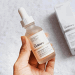 Hand holding The Ordinary Lactic Acid 10% + HA serum in a frosted glass dropper bottle with light pink liquid next to its product box.