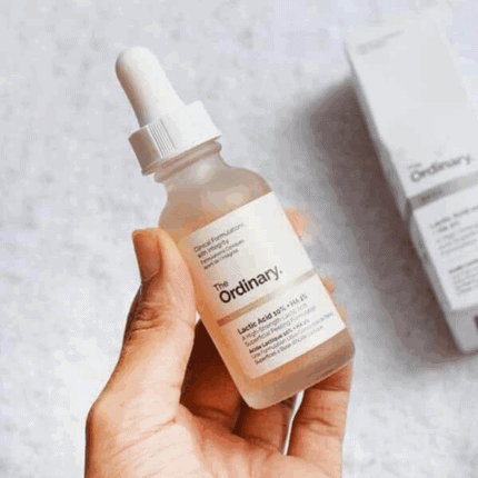 Hand holding The Ordinary Lactic Acid 10% + HA serum in a frosted glass dropper bottle with light pink liquid next to its product box.