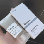 Hand holding The Ordinary Natural Moisturizing Factors + HA 30ml grey moisturizer tube alongside its white product box.