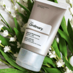 The Ordinary Natural Moisturizing Factors + HA 30ml grey moisturizer tube placed among green leaves and small white flowers.