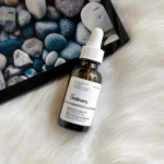 Flat lay of The Ordinary Retinol 0.5% in Squalane amber bottle placed on white fur surface next to tablet displaying pebble wallpaper.