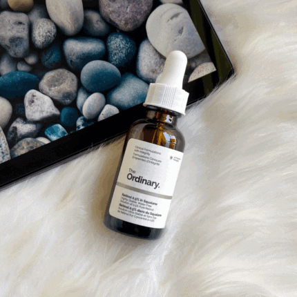 Flat lay of The Ordinary Retinol 0.5% in Squalane amber bottle placed on white fur surface next to tablet displaying pebble wallpaper.