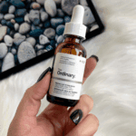 Person holding The Ordinary Retinol 0.5% in Squalane skincare bottle with white dropper against soft white textured background.