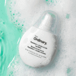 The Ordinary Squalane + Amino Acids Lip Balm 15ml in a white teardrop-shaped bottle placed among water and foam bubbles with a fresh aqua background.