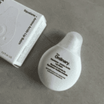 The Ordinary Squalane + Amino Acids Lip Balm 15ml in a white teardrop-shaped bottle placed on a grey surface next to its white product box.