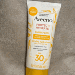 Single Aveeno Protect + Hydrate SPF 30 sunscreen tube placed upright on a soft, neutral-colored fabric background, showing the front label with broad-spectrum UVA/UVB protection and 3.0 fl oz (88 mL) size.