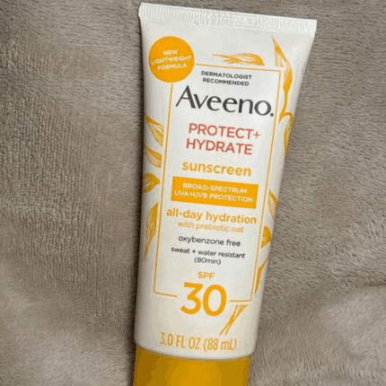 Single Aveeno Protect + Hydrate SPF 30 sunscreen tube placed upright on a soft, neutral-colored fabric background, showing the front label with broad-spectrum UVA/UVB protection and 3.0 fl oz (88 mL) size.