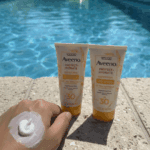 Two Aveeno Protect + Hydrate SPF 30 sunscreen tubes standing on a poolside ledge with blue water in the background, while a hand in the foreground shows a dollop of white sunscreen applied on the back of the skin.