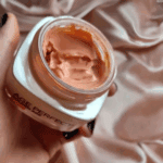 Close-up of a hand holding an open jar of Age Perfect Golden Age SPF 20 face cream with visible creamy texture, placed against a soft satin fabric background for a luxury skincare feel