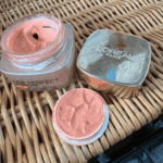 Opened Age Perfect Golden Age SPF 20 face cream jar with visible creamy texture, placed on a woven wicker surface alongside its lid and a small amount of cream in the cap, showing a real-life skincare setup