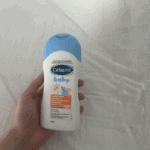 “Hand holding Cetaphil Baby moisturizing bath and wash bottle with blue cap on white fabric background, gentle baby cleanser”