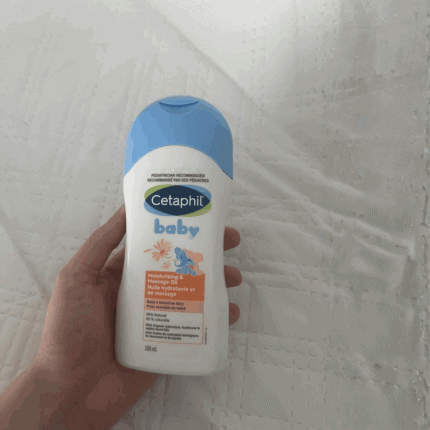 “Hand holding Cetaphil Baby moisturizing bath and wash bottle with blue cap on white fabric background, gentle baby cleanser”