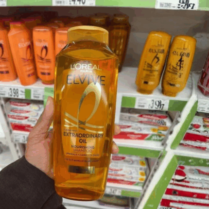 Hand holding L’Oréal Elvive Extraordinary Oil nourishing shampoo bottle in a retail store aisle with hair care products on shelves in the background, showcasing a product for dry and dull hair