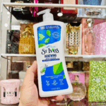 Hand holding St. Ives Renewing Collagen and Elastin body lotion pump bottle in front of a shelf with perfumes, showcasing a moisturizing skincare product in a real-life setting
